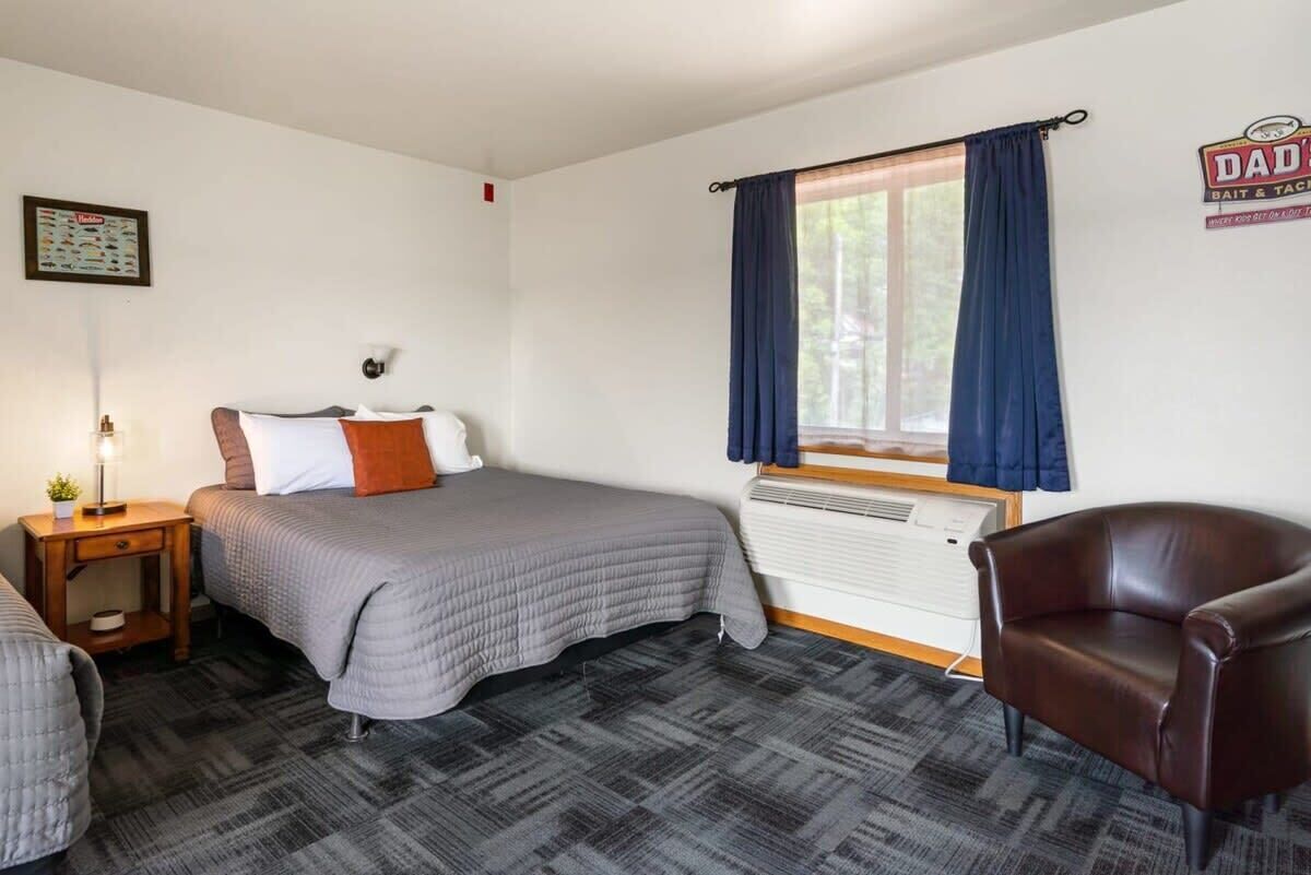 Une chambre avec deux lits, une chaise, une table de chevet, une lampe, une fenêtre avec des rideaux et un tableau ou une affiche au mur.