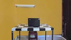 A small table with a laptop, a checkered tablecloth, and a wall-mounted shelf against a yellow wall. - SPOT ON 92475 Rc Homestay Syariah (Banyuwangi)