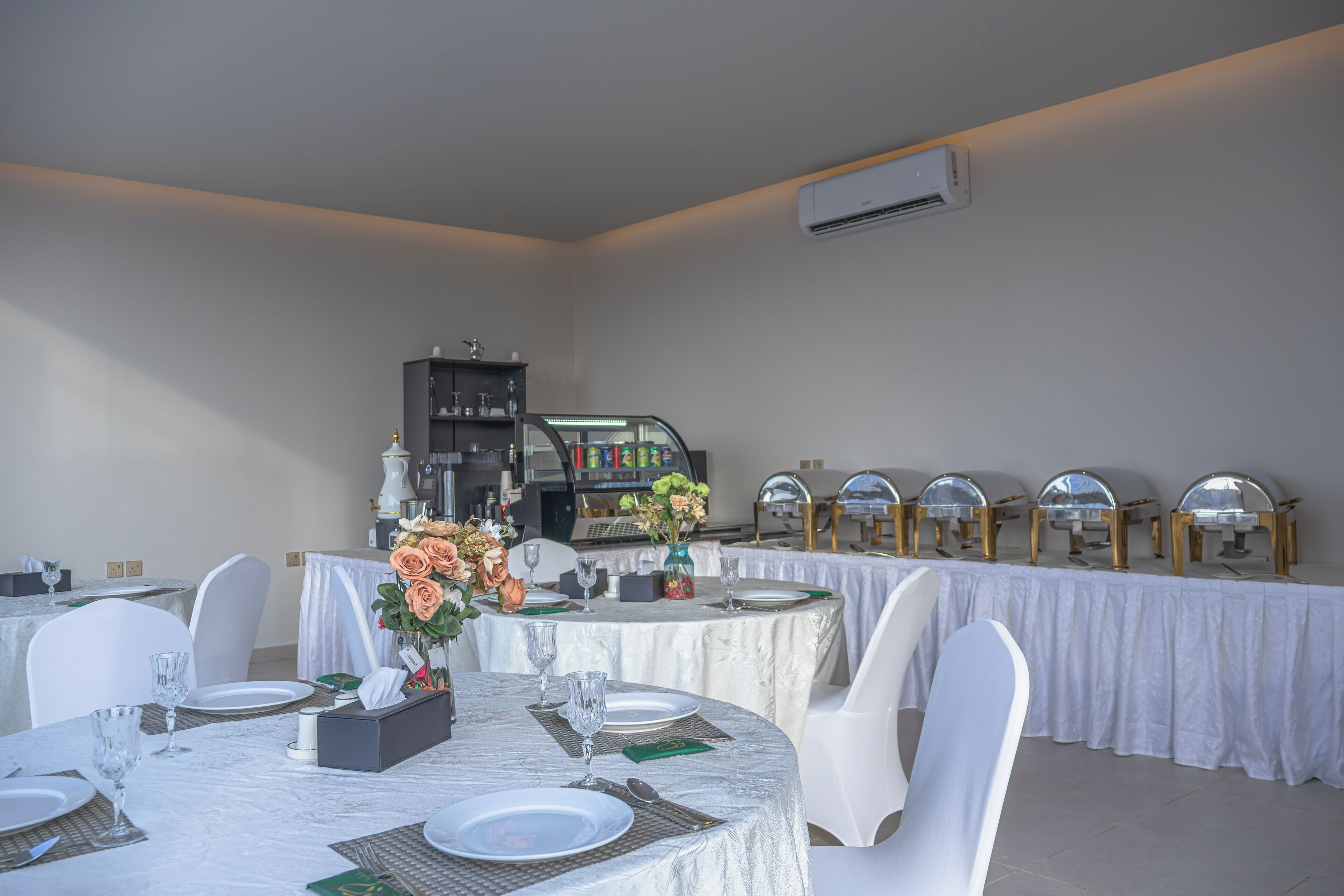 A dining area with round tables set for four, white chairs, and a buffet line with golden serving stations.