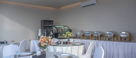 A dining area with round tables set for four, white chairs, and a buffet line with golden serving stations.