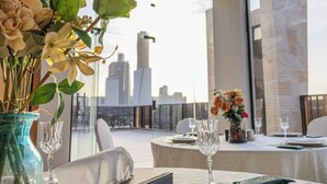 A dining table set for two with a vase of flowers, a glass, and a cityscape view in the background. - Royal Palace Hotel - Financial District (Riyadh)
