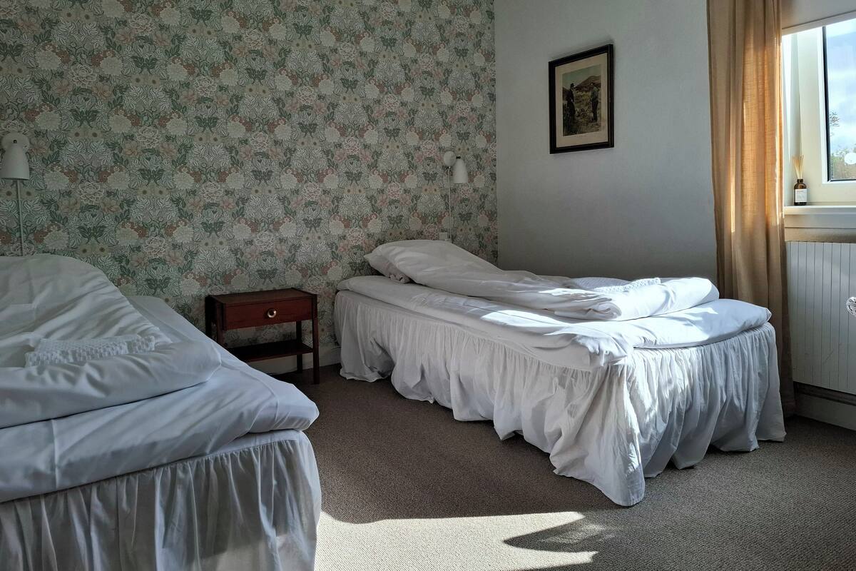 A hotel room with two beds, floral wallpaper, a framed picture on the wall, a nightstand, and a window with curtains.
