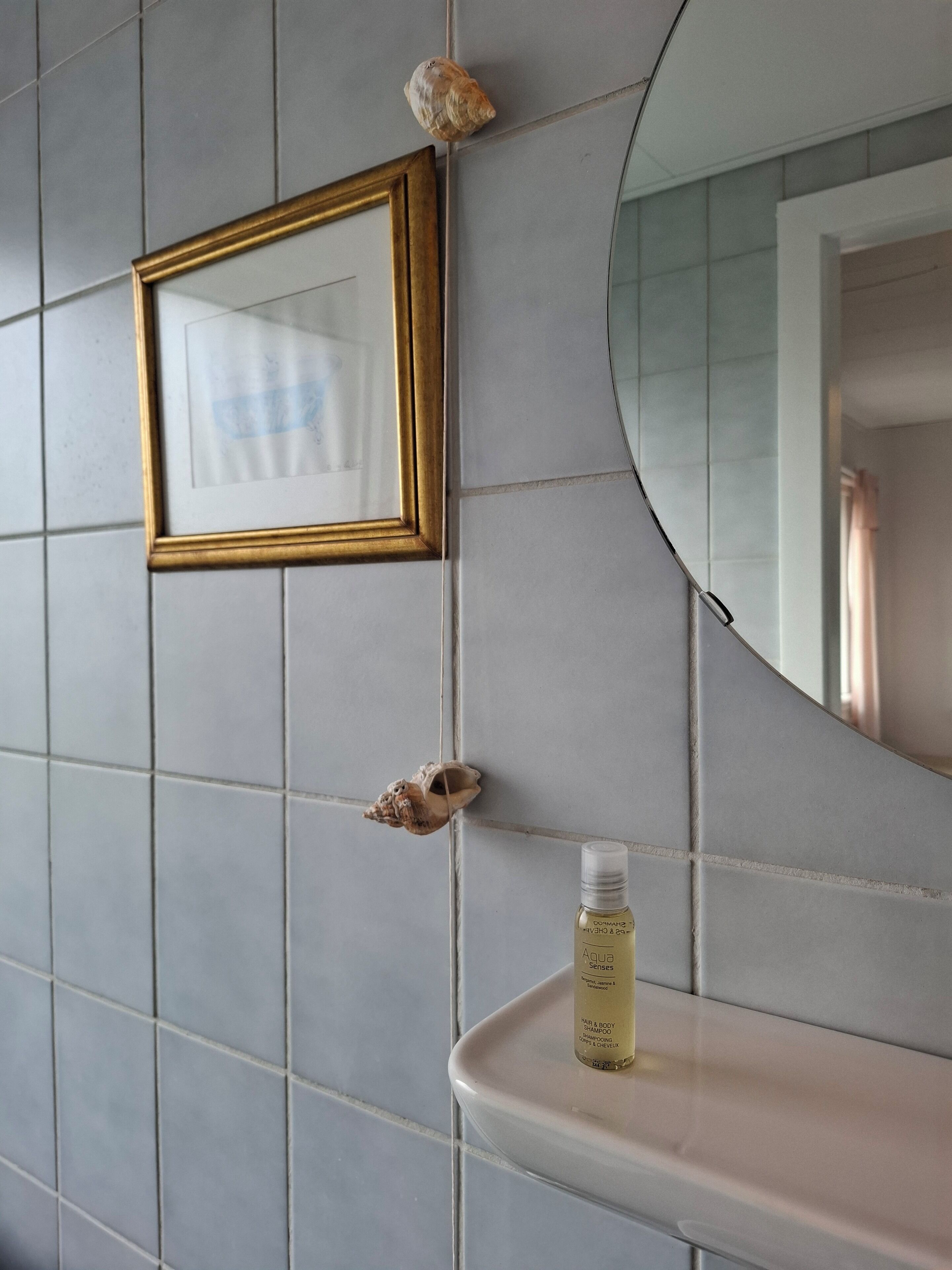 A bathroom with a tiled wall, a framed picture, a mirror, and a bottle of shampoo on a shelf.