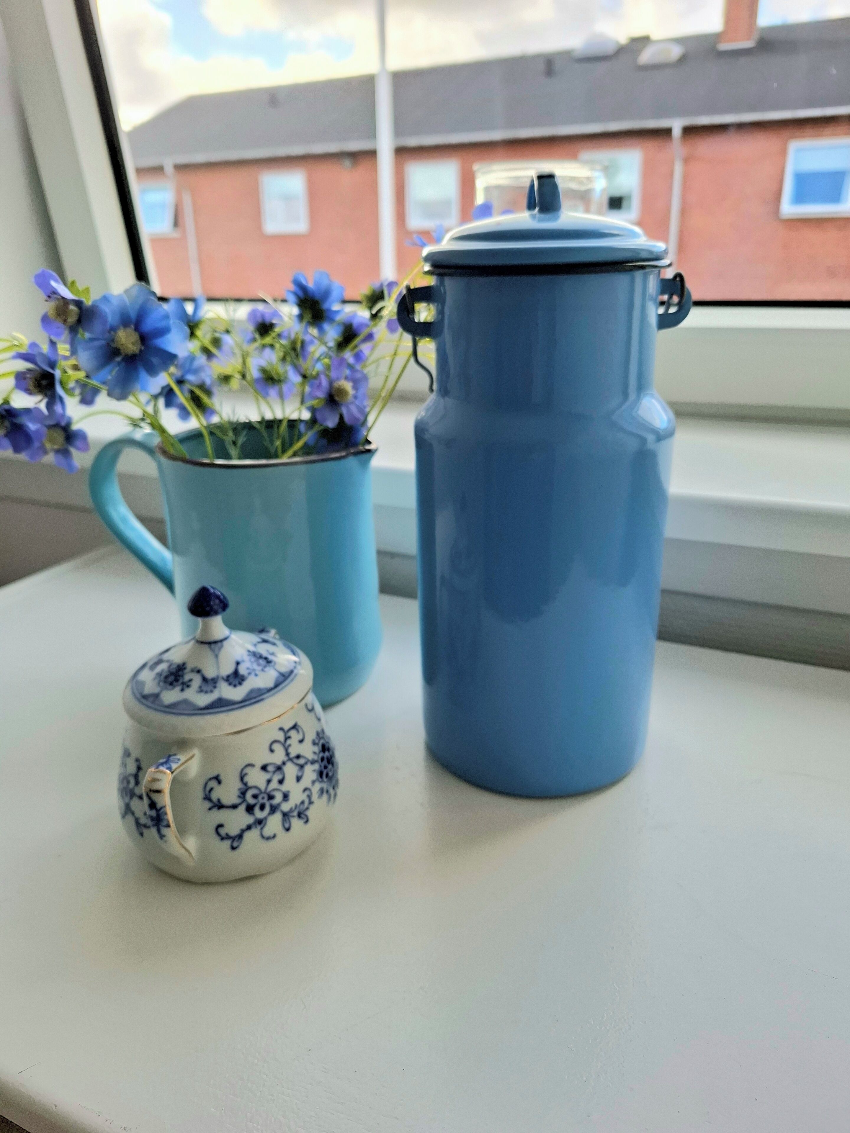 A window sill with a blue teapot, a blue mug with flowers, and a small blue teapot.
