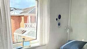 Una habitación pequeña con cama individual, ventana, cortinas blancas, radiador y un aplique de pared.