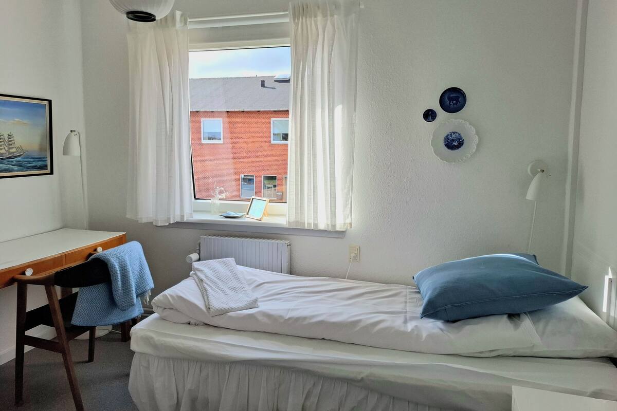 A bedroom with a single bed, a desk, and a chair. There is a window with curtains, a radiator, and a wall-mounted lamp.