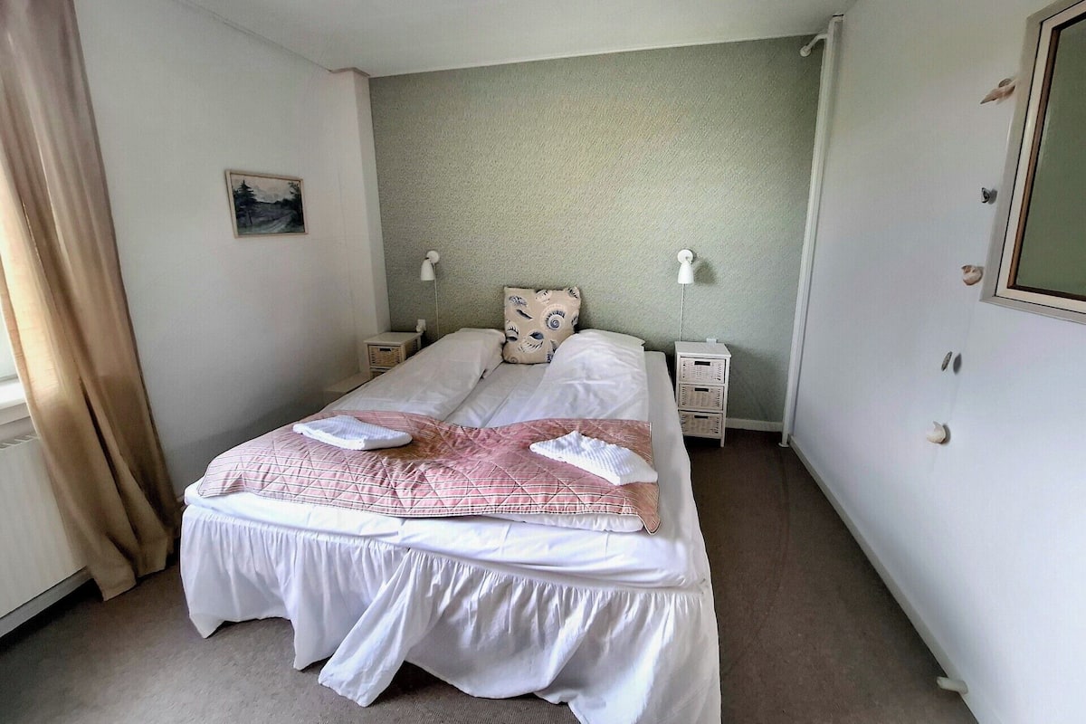 A double bed with a white and pink bedspread, two bedside lamps, a nightstand, and a window with curtains.