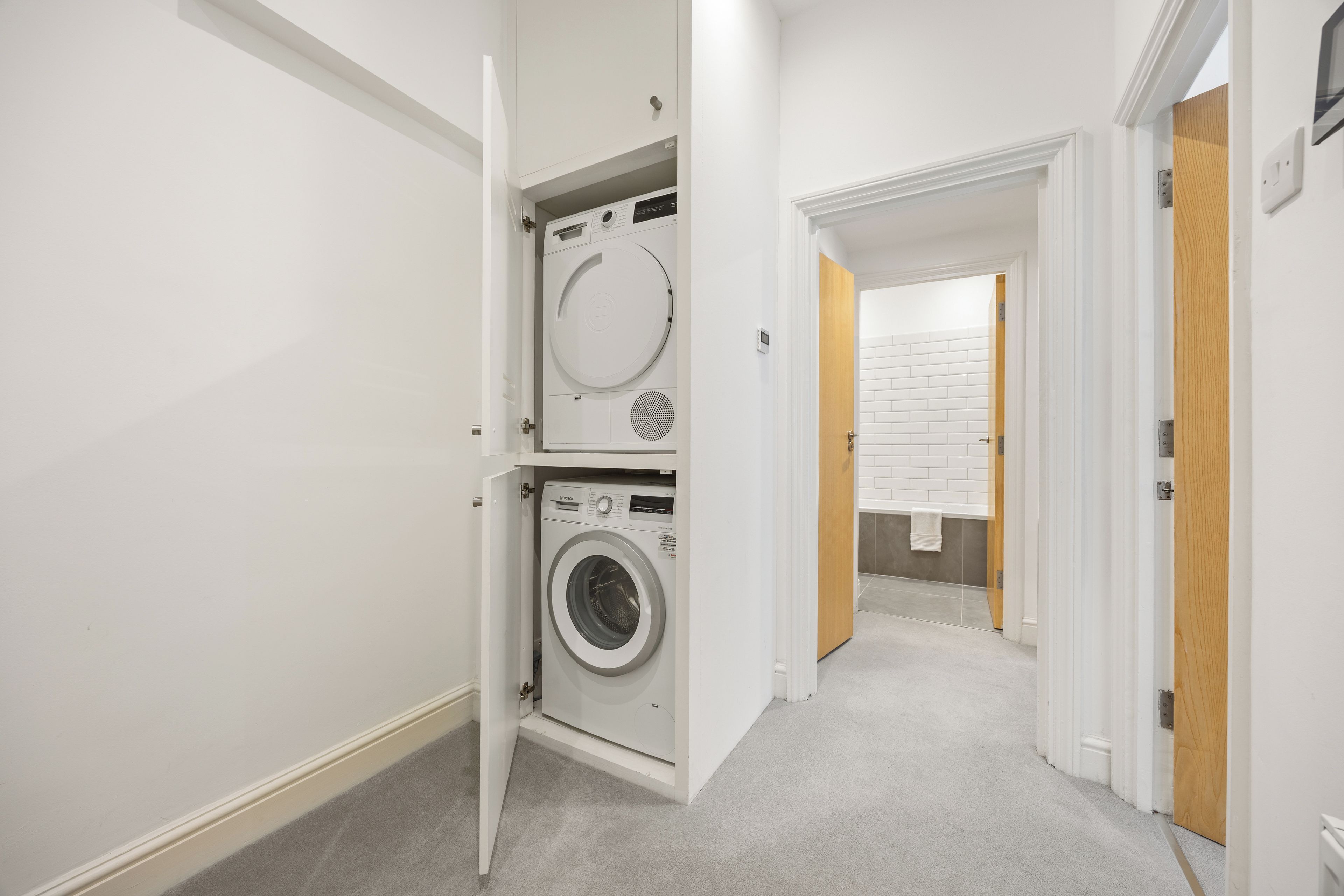 A laundry room with a front-loading washing machine and dryer, a white tiled wall, and a wooden door leading to a bathroom.