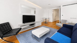 Ein modernes Wohnzimmer mit einem Flachbildfernseher, einer schwarzen Ledersofa, einem blauen Sofa, einem weißen Couchtisch und einem Essbereich mit einem Glastisch und Stühlen.