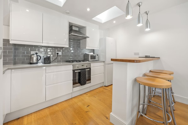A modern kitchen with white cabinetry, a gas stove, a microwave, and a kettle. There are three bar stools with metal legs.