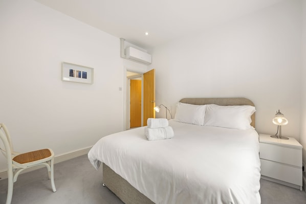 Un dormitorio con cama, mesita de noche, una silla y un cuadro en la pared.