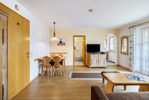 Una sala de estar con un sofá, una mesa de comedor de madera, sillas, un televisor y una zona de cocina con armarios y un radiador.