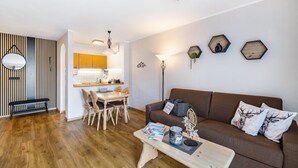 Una sala moderna con un sofá marrón, una mesa de comedor de madera y sillas. Hay un banco, una mesa de centro con revistas y una lámpara. Se ve la cocina al fondo, con armarios de madera y una encimera.