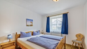 Un dormitorio con cama de madera, una mesita de noche, una silla, un ventanal con cortinas azules y un cuadro en la pared.