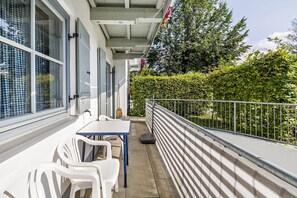 A balcony with a table and chairs, a broom, and a hedge in the background.