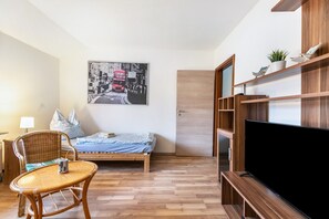 A room with a bed, a television, a chair, a table, and a wall-mounted shelf with a plant.