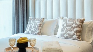 Un lit bien fait, agrémenté d’oreillers à motifs, une serviette pliée et deux verres, le tout posé sur un plateau avec un panier de fruits.