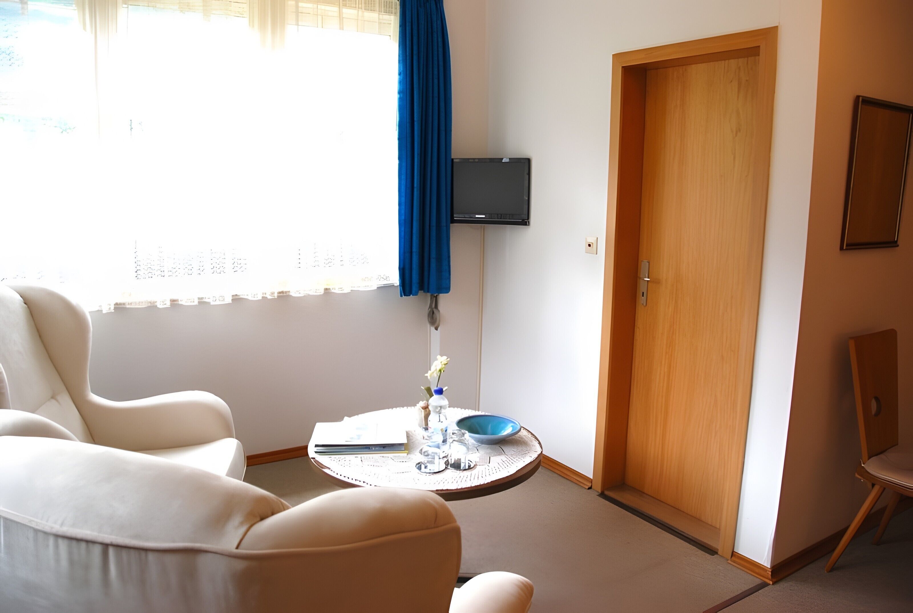Ein Zimmer mit Holztür, einem an der Wand befestigten Fernseher, einem runden Tisch mit Schüssel und Vase sowie zwei Stühlen.