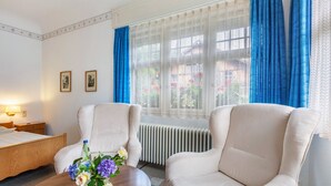 A hotel room with a bed, two armchairs, a round coffee table with flowers, a radiator, and a window with blue curtains. - Double Room No. 2 with Shared Terrace, Shared Garden and Wi-Fi (Dachsberg (Südschwarzwald))