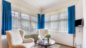 A room with two beige armchairs, a round table with a vase and bottles, a television, and large windows with blue curtains. - Double Room No. 2 with Shared Terrace, Shared Garden and Wi-Fi (Dachsberg (Südschwarzwald))