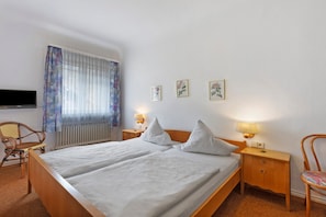 A double bed with two pillows, a wooden nightstand with a lamp, a chair, a TV, and three framed pictures on the wall.