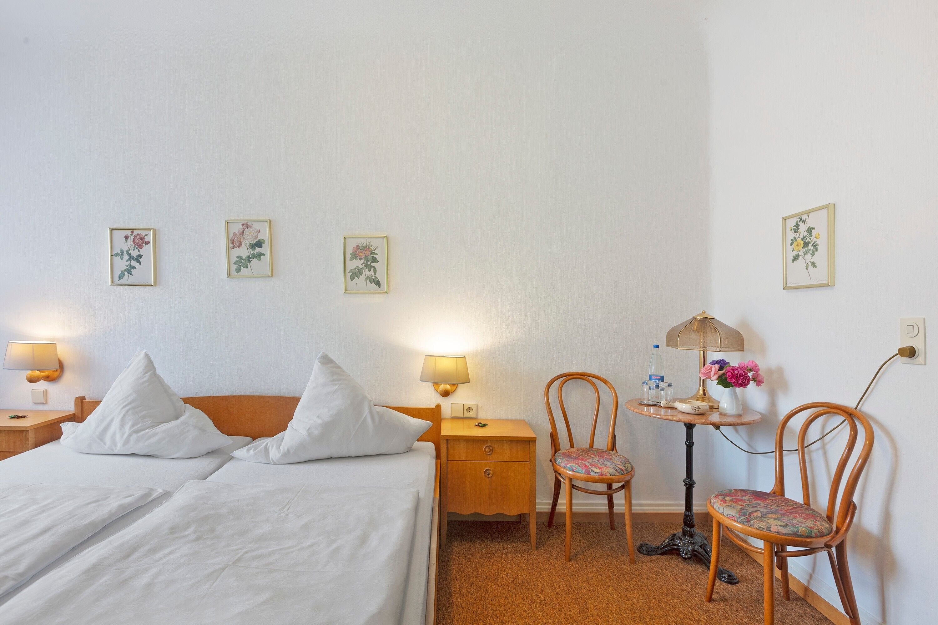 Ein Hotelzimmer mit einem Bett, zwei Nachttischen mit Lampen, einem Sessel und einem kleinen Tisch ebenfalls mit einer Lampe, sowie Wandbildern mit floralem Dekor.