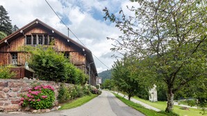 A rustic wooden building with a stone foundation, a paved road, and a grassy area with a tree. - Double Room No. 4 with Mountain View, Shared Terrace and Wi-Fi (Dachsberg (Südschwarzwald))