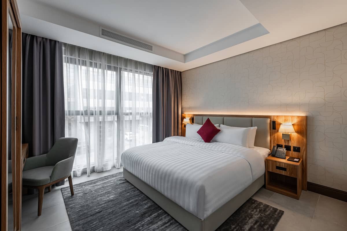 Habitación de hotel con una cama grande, una silla, una mesita de noche con lámpara y vistas a la ciudad a través de cortinas translúcidas.