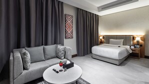 A hotel room with a grey sofa, a round coffee table with a remote control, a wine glass, and a small plant. There is a bed with a white comforter and a bedside table with a lamp. - Lafait Hotel Serviced Apartments (Al Khobar)