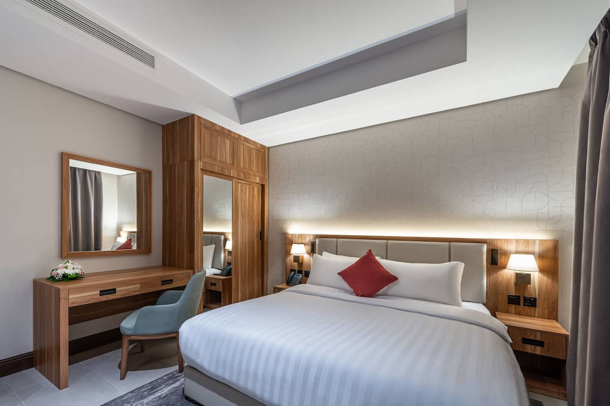 Habitación de hotel con una cama grande, un escritorio de madera, una silla, una lámpara de escritorio, una mesita de noche con lámpara y un espejo en la pared.