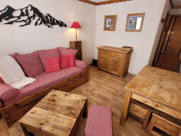 Una sala acogedora con un sofá a cuadros rojo y blanco, una mesa de centro de madera, una mesa de comedor de madera, un mueble pequeño y una pintura mural con la silueta de montañas.