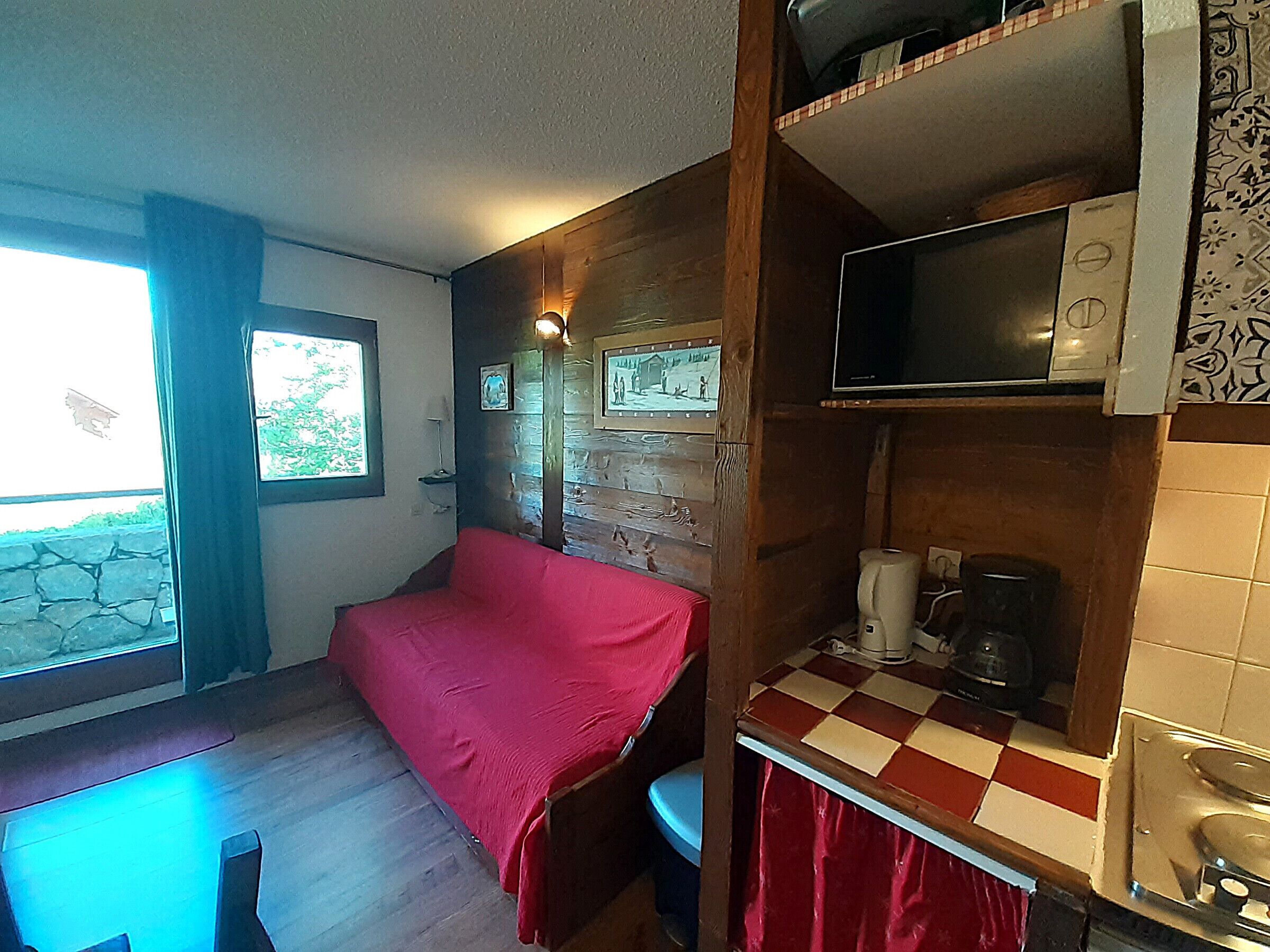 Une pièce de taille modeste comprenant un lit, une petite table, une fenêtre avec des rideaux, un téléviseur fixé au mur et un coin cuisine.