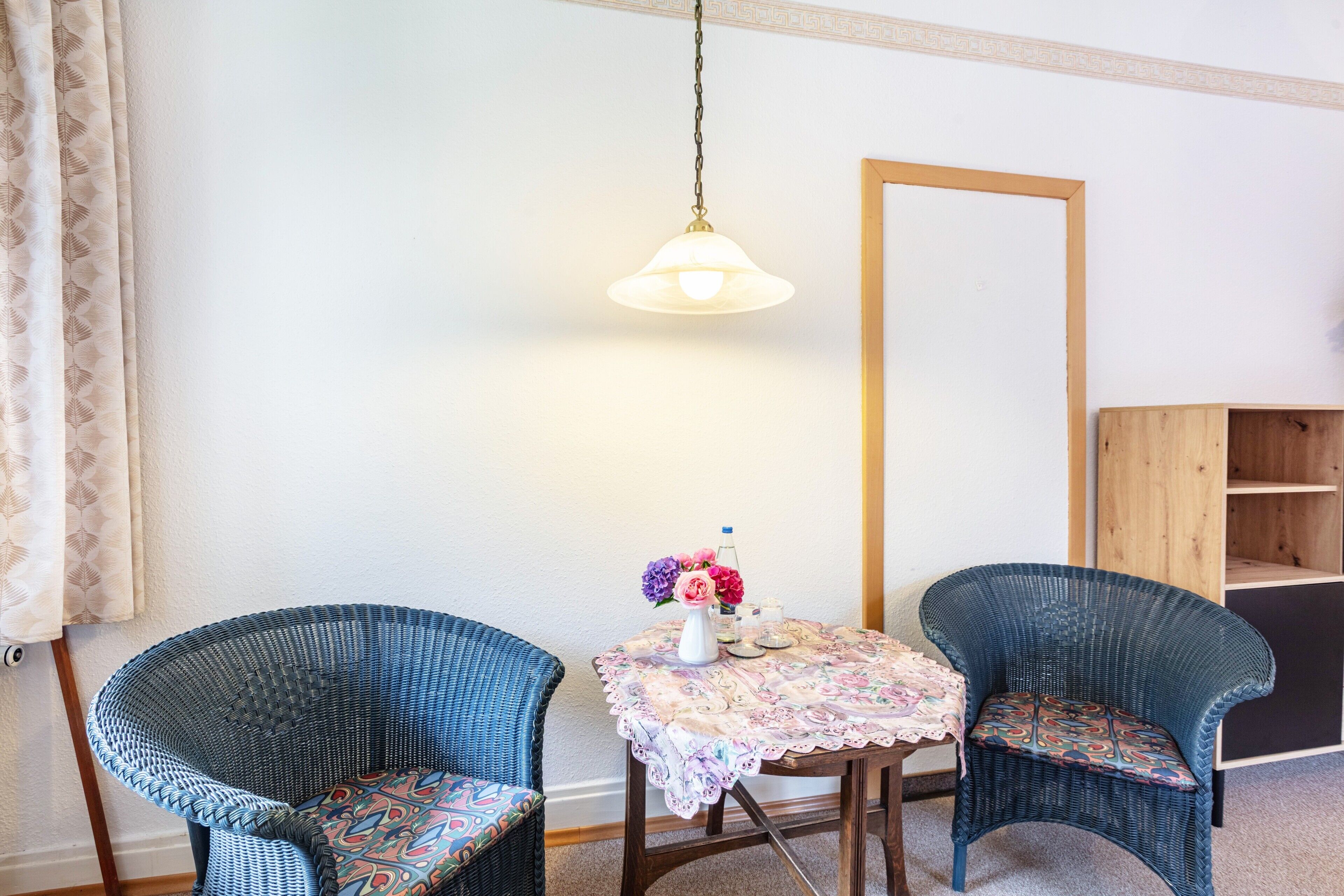 A room with two wicker chairs, a small table with a lace tablecloth, a hanging light fixture, and a wooden cabinet.