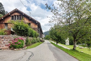 A rustic wooden building with a stone foundation, a paved road, and a grassy area with a tree.