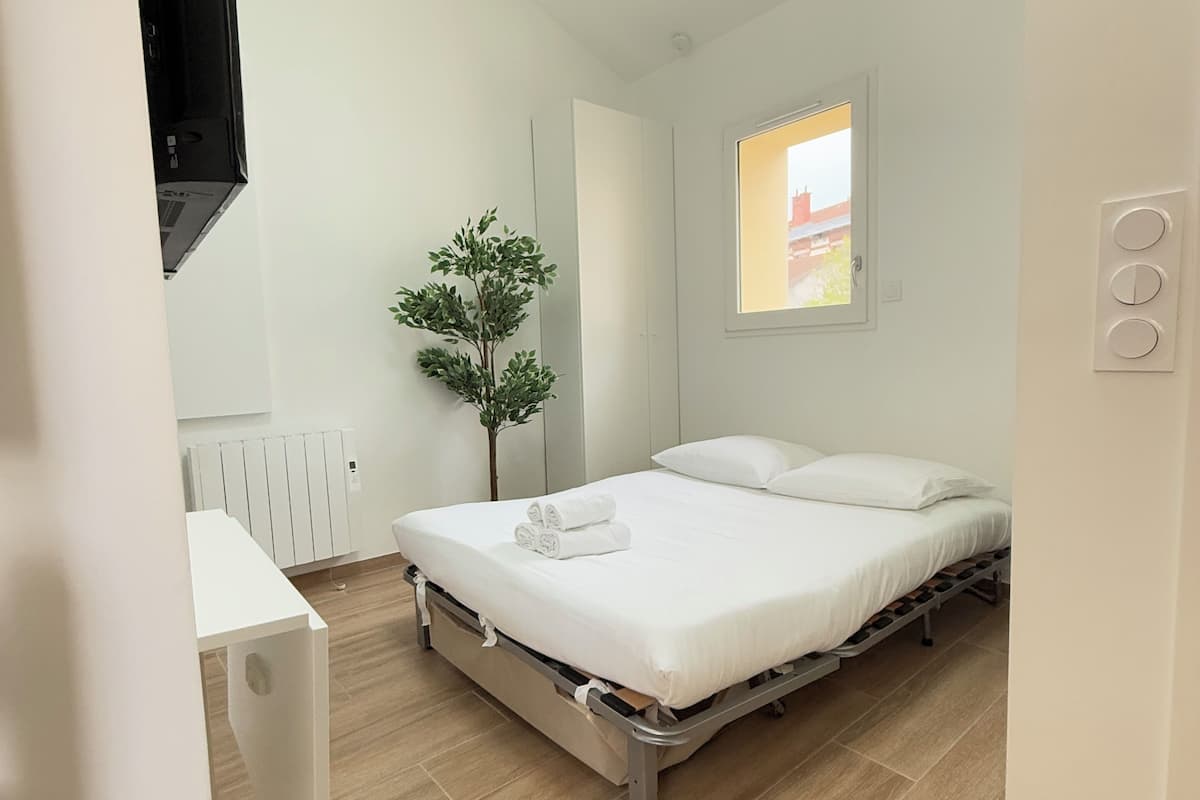 A modern bedroom with a bed, a small potted plant, a window with a view, and a white desk with a flat-screen TV.