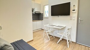 Ein modernes Wohnzimmer mit einer Couch, einem Esstisch mit Stühlen, einem an der Wand montierten Fernseher und einer Küche im Hintergrund.