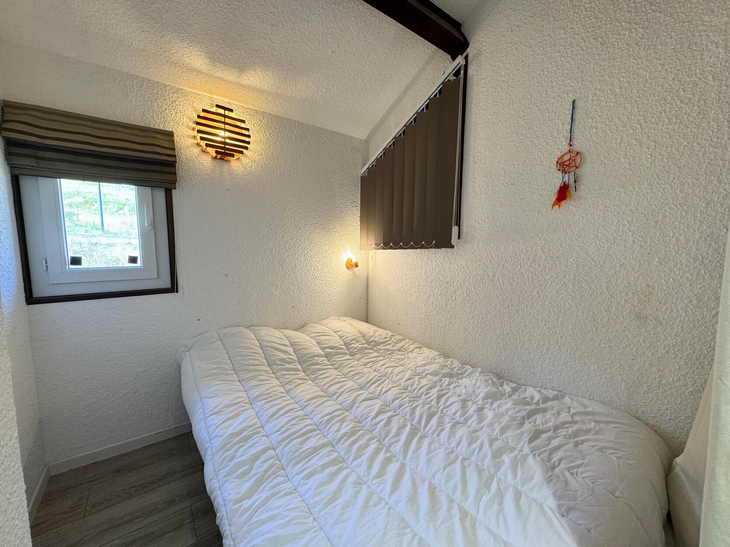 A bedroom with a single bed, a window with blinds, a wall-mounted light fixture, and a dreamcatcher decoration.