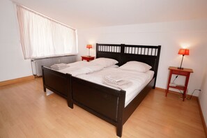 Ein Doppelbett mit schwarzem Kopfteil, zwei Nachttische jeweils mit einer Lampe, eine Heizung und ein Fenster mit durchsichtigen Vorhängen.