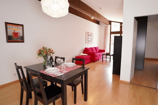Ein Essbereich mit einem Holztisch, Stühlen, einer Blumenvase und einer roten Couch im Hintergrund.