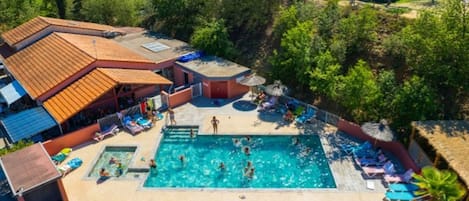 Un complexe hôtelier doté d’une piscine centrale entourée de chaises longues et de parasols, d’un espace couvert avec un toit en tuiles et d’un décor naturel composé d’une forêt en arrière-plan.