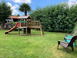A wooden play structure with a slide and climbing area, surrounded by a grassy yard and a hedge.