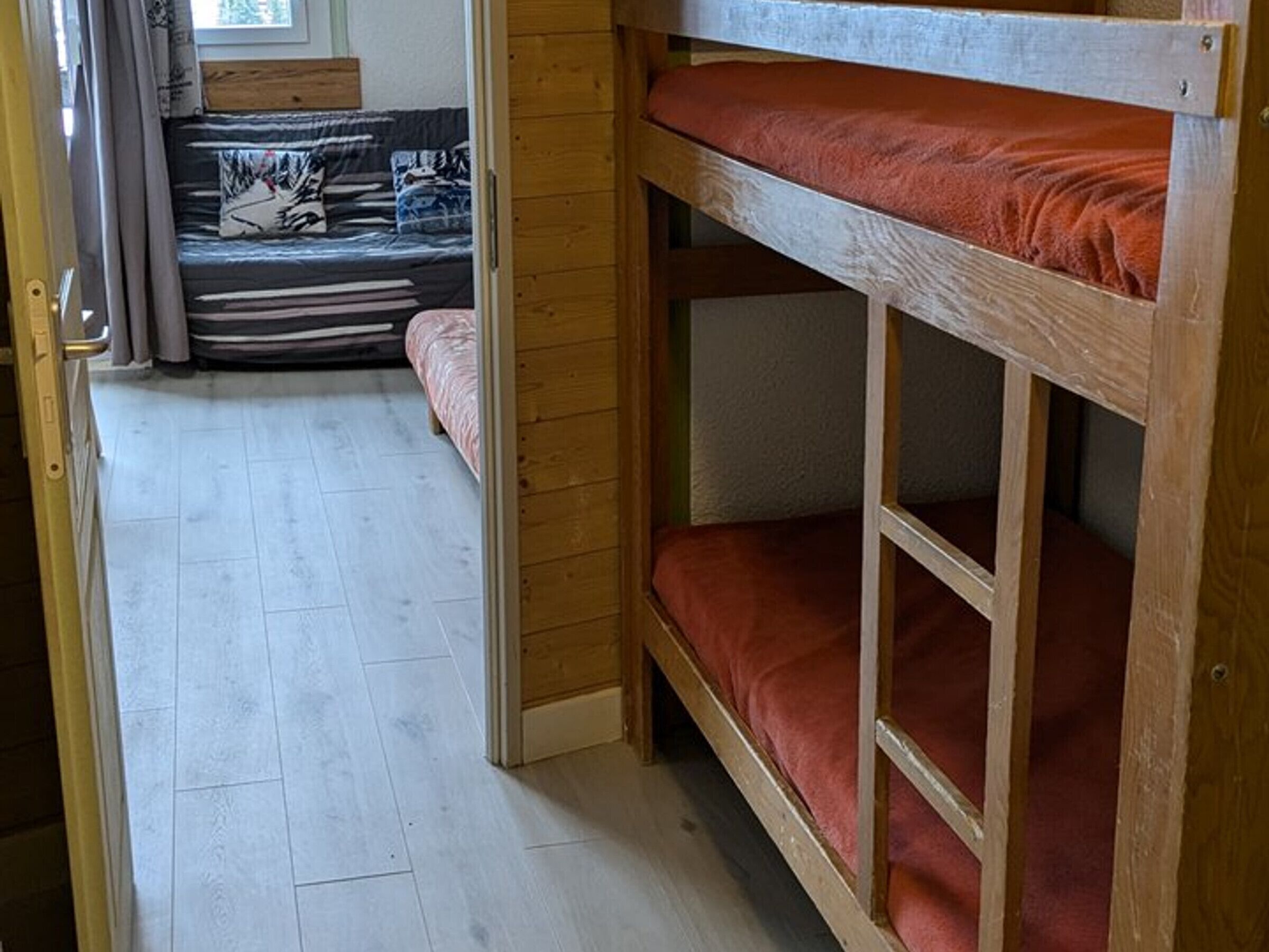 A bunk bed with a red mattress, a wooden floor, and a sofa with patterned cushions in the background.