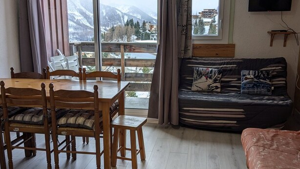 A room with a dining table and chairs, a sofa with patterned cushions, a television mounted on the wall, and a view of snow-covered mountains through large windows. - Maxi studio 6 pers, central Orcières, near slopes (Orcières Merlette)