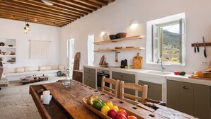 A rustic kitchen with wooden beams, a dining table with fruit, and a view of a mountainous landscape through the window.