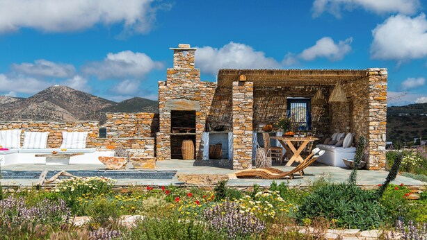 A stone building with a thatched roof, a seating area with cushions, and a table with chairs.