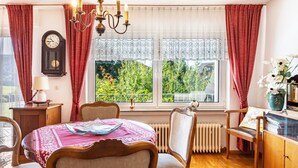A dining area with a table set for four, a clock on the wall, and a view of greenery outside. - Apartment 'Kropp' with Mountain View, Balcony and Wi-Fi (Steinen)