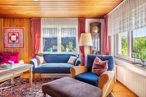 A cozy living room with a blue sofa, a wicker chair, a table with a lamp, and a view of greenery outside. - Apartment 'Kropp' with Mountain View, Balcony, and Wi-Fi (Steinen)