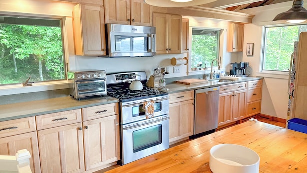 A modern kitchen with wooden cabinets, stainless steel appliances, and large windows offering a view of the outdoors.