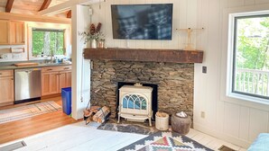 A rustic interior with a stone fireplace, wooden beams, a vintage radio, and a large window overlooking greenery.
