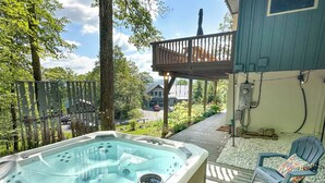 A hot tub on a wooden deck with chairs, surrounded by greenery and a house in the background.